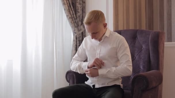 Handsome groom man dresses in wedding morning and fixes his buttons on a white shirt sleeve, indoors — Stockvideo