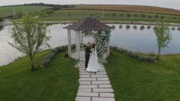 Pasgetrouwden, bruidegom, bruid omarmen, knuffelen, kussen, bruiloft huwelijksceremonie, boog met bloemen — Stockvideo