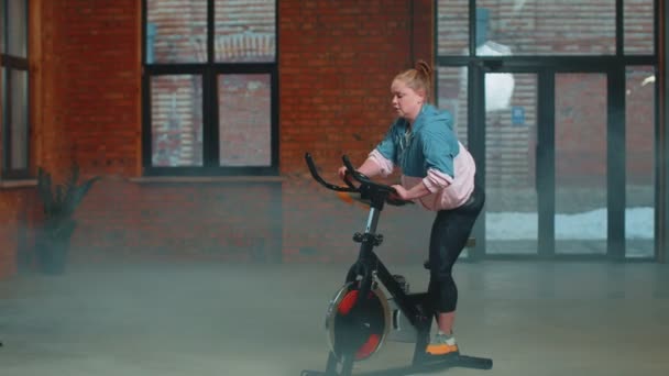 Atletická dívka provádějící aerobní jízdu na koni cvičení na kole stacionární kolo v mlhavé tělocvičně — Stock video
