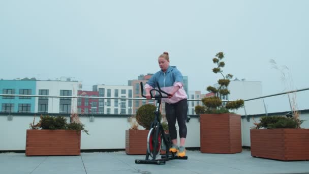 Здорова кавказька жінка тренується на стаціонарному велосипеді на даху будинку. — стокове відео