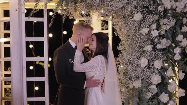 Nygifta, brudgum, brud omfamning, kramar, kyssar, bröllop kväll ceremoni, båge med blommor — Stockvideo