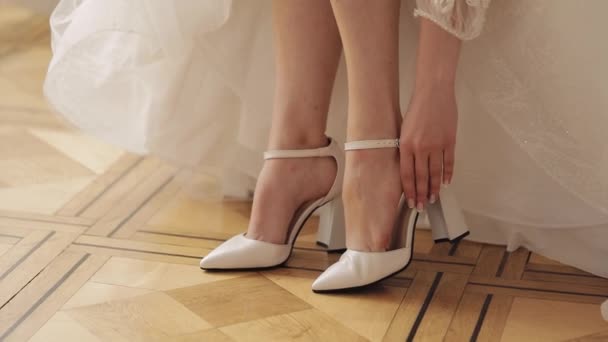 Junge Braut im weißen Hochzeitskleid trägt hochhackige Schuhe an schlanken Beinen, während sie zu Hause sitzt — Stockvideo