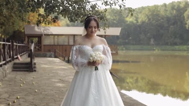 Mooie stijlvolle bruid in witte trouwjurk en sluier houden bruiloft boeket in handen in park — Stockvideo