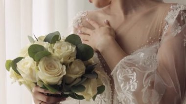 Beyaz elbiseli gelin çiçek buketli pencerenin yanında kalıyor, düğün hazırlıklarını evde yapıyor.