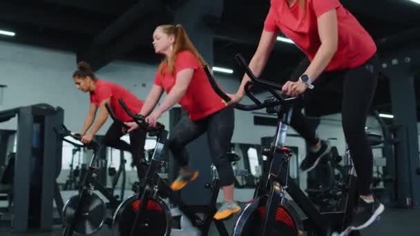 Mosolygós barátok csoportja női osztály gyakorlása, edzés, forgó álló kerékpár a modern tornaterem — Stock videók