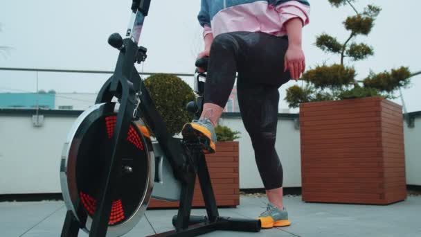 Athletic girl performing aerobic riding training exercises on cycling stationary bike on house roof — Stockvideo