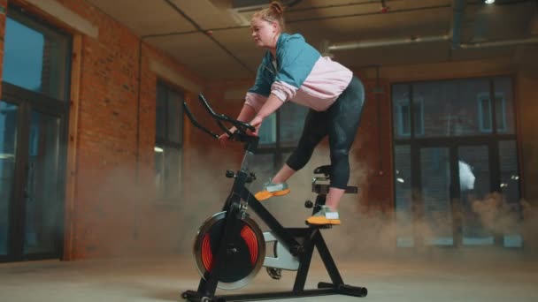 Athletic girl performing aerobic riding training exercises on cycling stationary bike in foggy gym — Stock Video