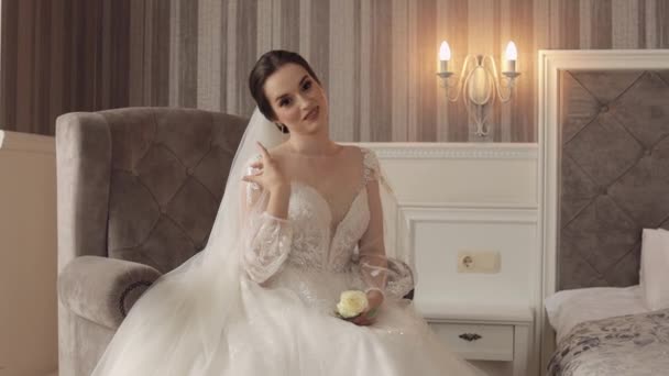 Κορίτσι νύφη σε λευκό φόρεμα κάθεται στην καρέκλα στο υπνοδωμάτιο σπίτι με λουλούδια γαμήλιο μπουκέτο για τον γαμπρό — Αρχείο Βίντεο