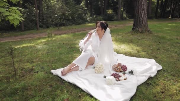 Mariée en robe de boudoir assis dans le jardin du matin buvant champagne femme en robe de nuit voile — Video