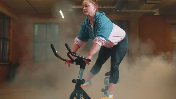 Athletic girl performing aerobic riding training exercises on cycling stationary bike in foggy gym — Vídeo de Stock