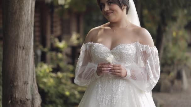 Красива стильна наречена в білій весільній сукні і завісі, що тримає весільний букет в руках в парку — стокове відео