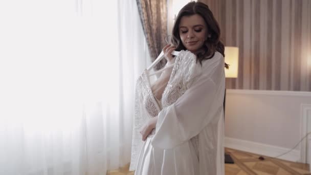 Bride in lingerie standing with her wedding dress at home near window, white boudoir dress and veil — Vídeo de Stock