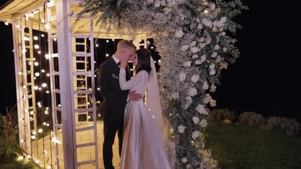 Friss házasok, vőlegény, menyasszony ölelés, ölelés, csók, esküvő esti szertartás, boltív virágokkal — Stock videók