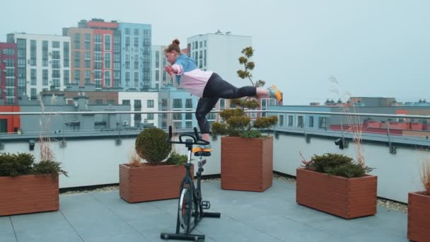 Athletic girl performing aerobic riding training exercises on cycling stationary bike on house roof — Stock Video