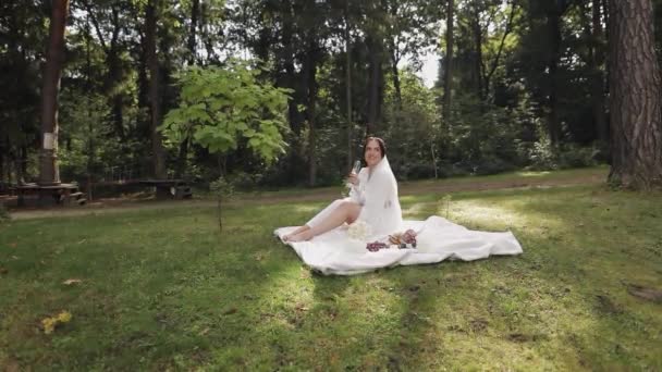 Весільна наречена в будуарній сукні, сидячи в ранковому саду, п'є шампанське жінку в нічній шубці — стокове відео