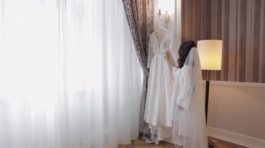 Gelin iç çamaşırlarıyla dans ediyor. Gelinliği pencerenin yanında, beyaz elbisesi ve duvağıyla.