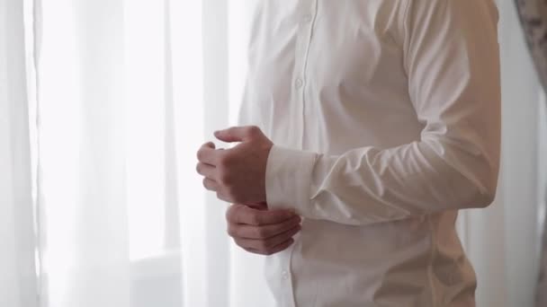 Όμορφος γαμπρός άντρας ντύνεται το πρωί του γάμου και βάζει τα κουμπιά του σε ένα λευκό πουκάμισο μανίκι, σε εσωτερικούς χώρους — Αρχείο Βίντεο