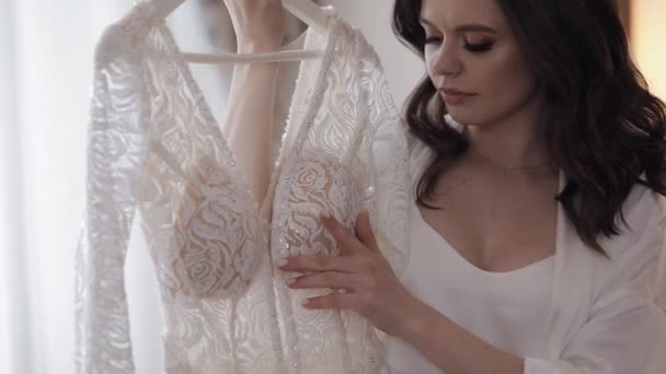 Noiva em lingerie de pé com seu vestido de noiva em casa perto da janela, vestido boudoir branco e véu — Vídeo de Stock