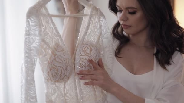 Sposa in lingerie in piedi con il suo abito da sposa a casa vicino alla finestra, vestito boudoir bianco e velo — Video Stock