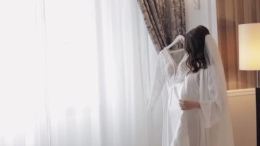 Gelin iç çamaşırlarıyla dans ediyor. Gelinliği pencerenin yanında, beyaz elbisesi ve duvağıyla.