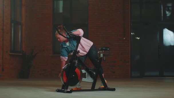 Athletic girl performing aerobic stretching training twine exercises on cycling stationary bike — 비디오