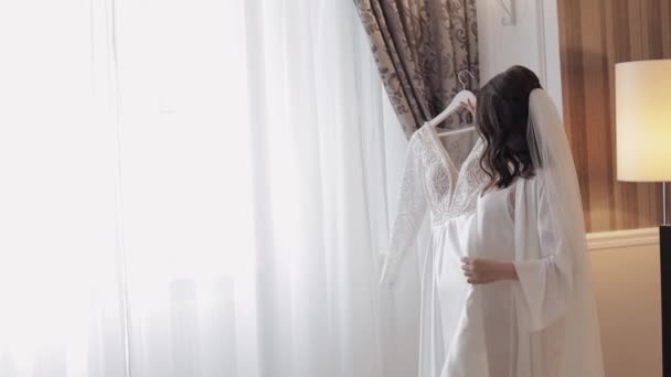 Νύφη με εσώρουχα χορεύει με το νυφικό της στο σπίτι κοντά στο παράθυρο, λευκό φόρεμα μπουντουάρ και πέπλο — Αρχείο Βίντεο