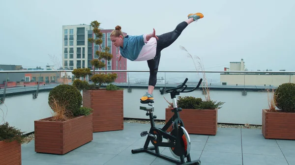 Athletic girl performing swallow acrobatic trick exercises on cycling stationary bike on house roof