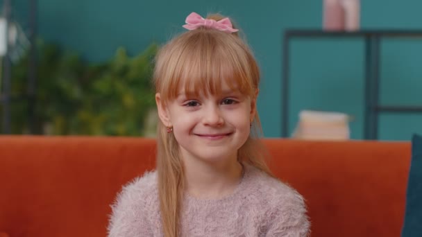 Porträt eines kleinen entzückenden Mädchens, das allein zu Hause auf dem Sofa sitzt, in die Kamera schaut und lächelt — Stockvideo