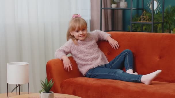 子供女の子横たわっソファに家でカメラを見て、笑顔、手ジェスチャーこんにちはまたはさようなら手を振って — ストック動画
