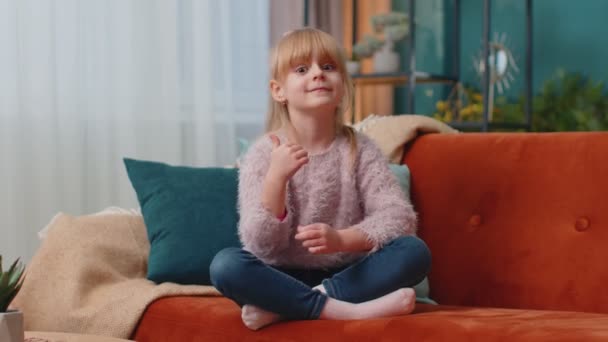 Anak perempuan duduk di sofa di rumah sendirian menunjukkan jempol seperti tanda positif sesuatu yang baik — Stok Video