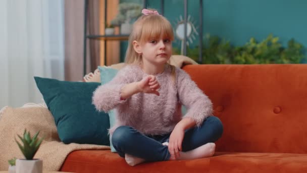 Dziecko dziewczynka dziecko siedzi na kanapie w domu sam pokazując kciuki w dół znak gest, niechęć, dezaprobata — Wideo stockowe