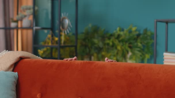 Kindermädchen spielt Versteckspiel in der Nähe des Sofas allein zu Hause, schaut in die Kamera und lächelt — Stockvideo