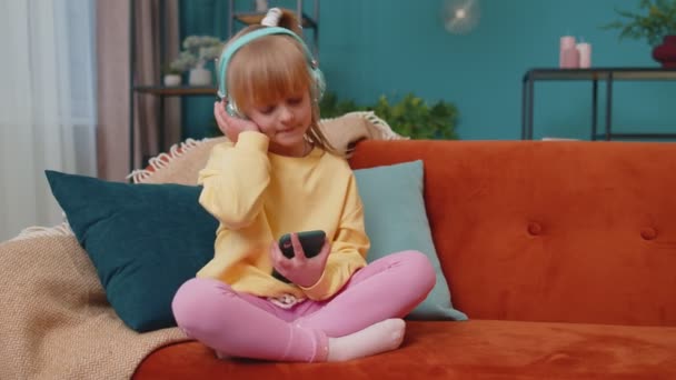 Kindermädchen mit Smartphone im Kopfhörer tanzen, während sie zu Hause allein auf dem Sofa Musik hören — Stockvideo