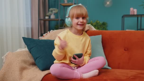 Niño niña con smartphone en auriculares bailando cantando escuchando música en casa solo en el sofá — Vídeo de stock