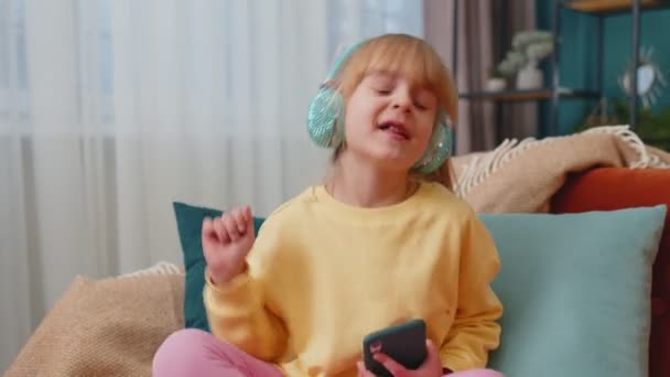 Niño niña con smartphone en auriculares bailando cantando escuchando música en casa solo en el sofá — Vídeo de stock