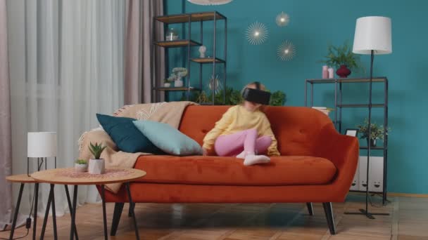 Тоддлер девушка сидит на домашнем диване, используя приложение шлем виртуальной реальности, чтобы играть в симуляцию игры — стоковое видео