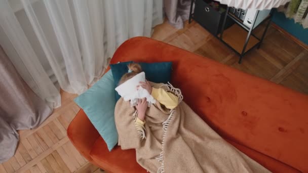 Дитяча дівчинка в плетених страждає від холоду або алергії, дме ніс в серветку на дивані вдома — стокове відео