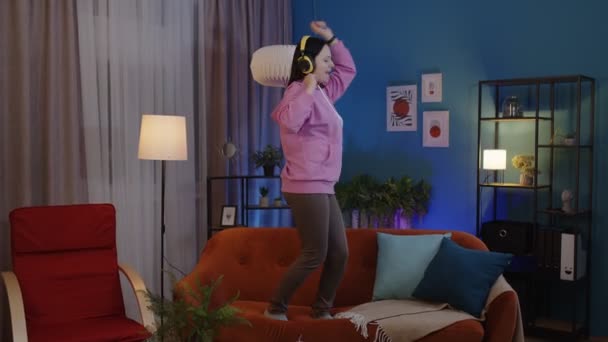 Щаслива доросла жінка в навушниках танцює і стрибає, слухаючи музику вдома наодинці на дивані — стокове відео