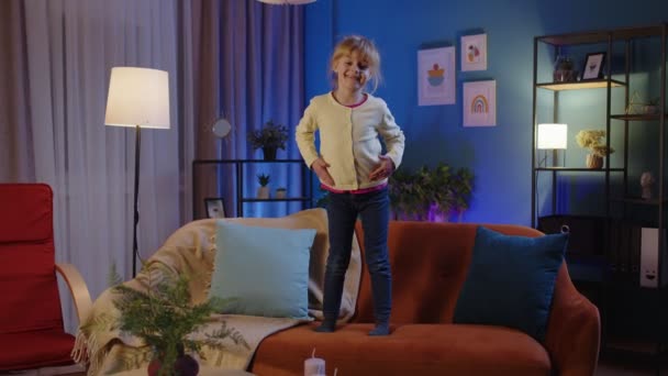Happy dziecko dziewczyna taniec modny taniec podczas słuchania muzyki w domu sam stojąc na kanapie — Wideo stockowe