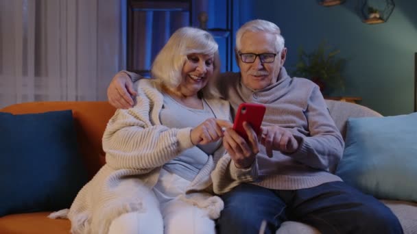 Szczęśliwa uśmiechnięta para seniorów ze smartfonem na kanapie w domu spędza czas wolny w sieciach społecznościowych — Wideo stockowe