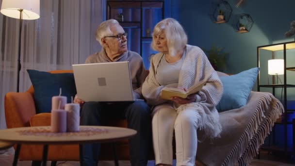 Senior παππούδες ζευγάρι ανάγνωση βιβλίο, χρησιμοποιώντας το φορητό υπολογιστή στον καναπέ στο σαλόνι νύχτα στο σπίτι — Αρχείο Βίντεο