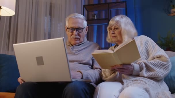 Senioren-Großelternpaar liest Buch, nutzt Laptop-PC auf Couch im heimischen Wohnzimmer — Stockvideo