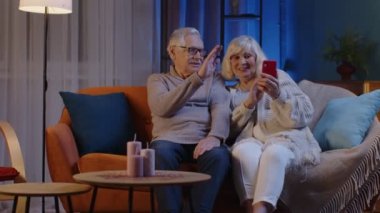 Gülümseyen yaşlı çift, büyükannem ve büyükbabam evdeki koltukta cep telefonuyla video görüşmesi yapıyor.