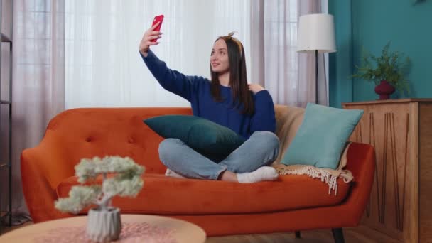 Mujer en el sofá en casa con teléfono inteligente tomando selfie en la cámara del teléfono móvil, videollamada virtual en línea — Vídeo de stock