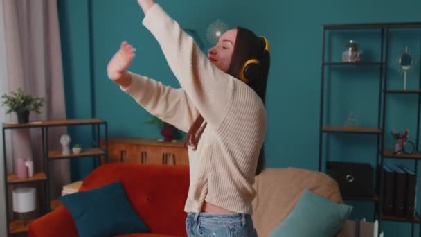 Kablosuz kulaklıklı neşeli genç kadın dans ediyor, evdeki oturma odasında rahat bir kanepede şarkı söylüyor. — Stok video