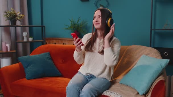 Überglückliche junge Frau mit kabellosen Kopfhörern tanzt, singt auf gemütlicher Couch im heimischen Wohnzimmer — Stockvideo
