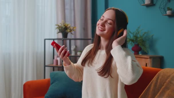 Mujer joven bastante caucásica en auriculares escuchando música bailando, cantando en la sala de estar en casa — Vídeos de Stock