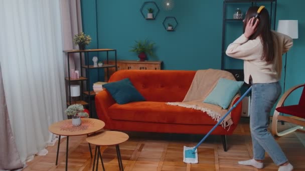 Joyful jovem mulher está limpando chão na sala de estar apartamento, se divertindo ouvindo música em casa — Vídeo de Stock