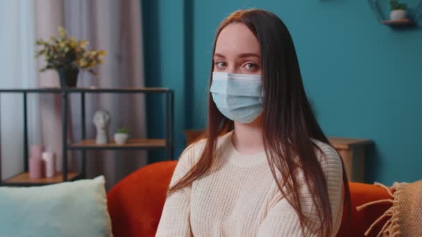 Портрет молодой больной девушки в защитной маске, сидящей в гостиной и смотрящей в камеру — стоковое видео
