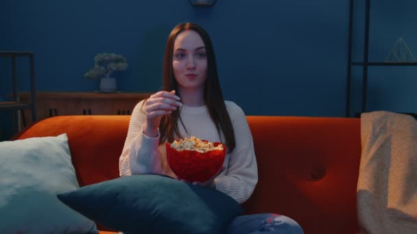 Wanita duduk di sofa sendirian, makan popcorn dan menonton serial TV yang menarik, film horor di rumah — Stok Video